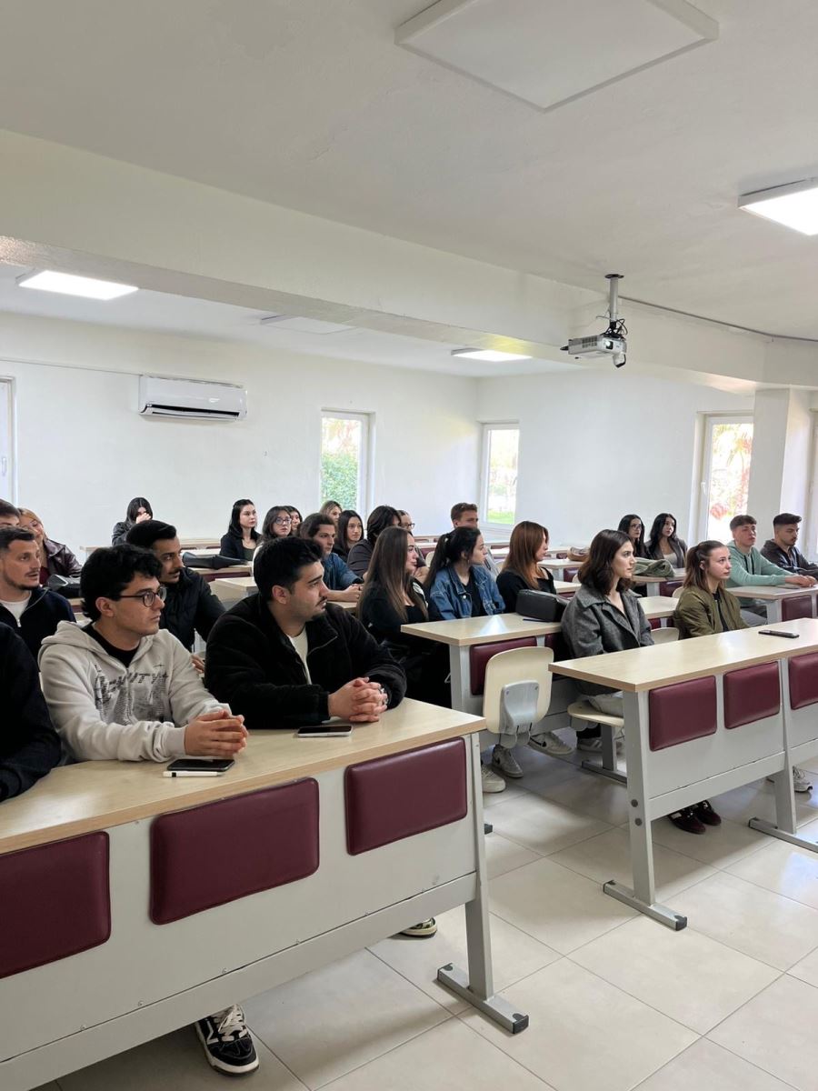 In a university classroom, students are seated at desks and listening to a presentation. They are facing the front of the room and attentively following the meeting. The classroom is bright and organized, and the students appear focused on the speaker and the ongoing discussion.